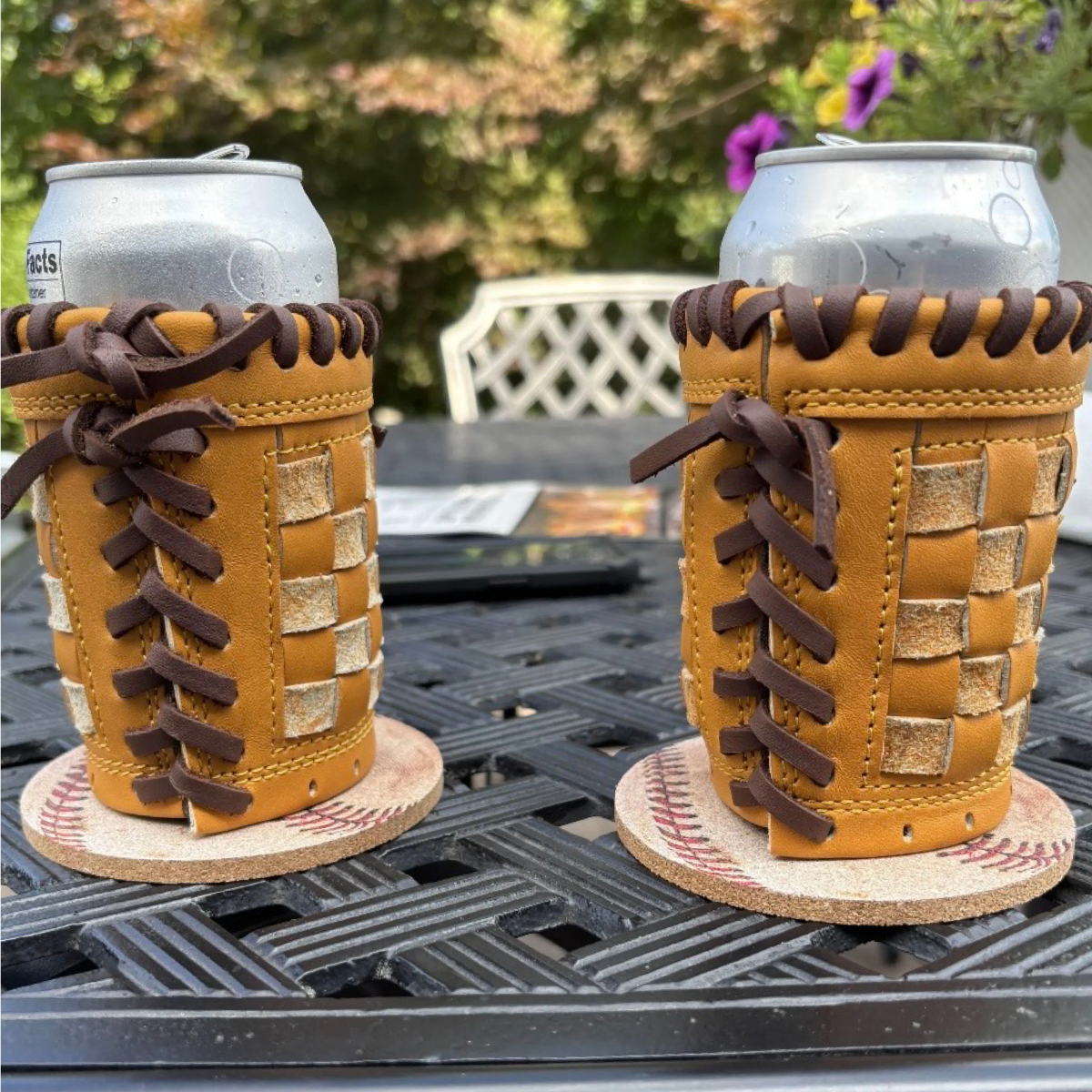 Two tan leather beverage can coolers with dark brown laces and woven tan accents, shown on a black outdoor table with cans inserted. The background shows a blurred garden and patio setting.